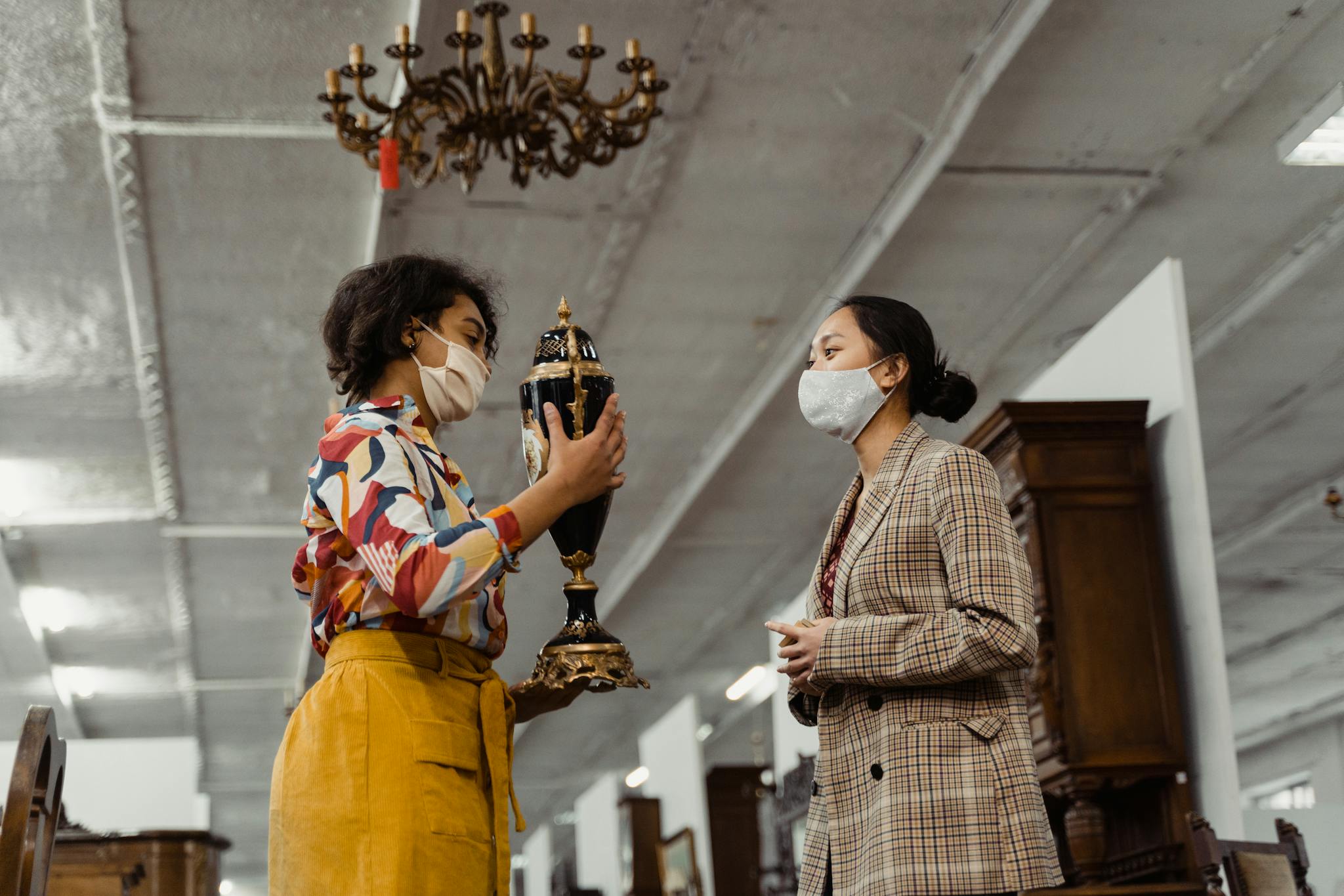 Two women in masks evaluating an antique vase in a shop, showcasing vintage fashion.